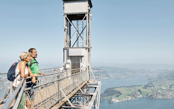Ein Mann und eine Frau stehen in Wanderkleidung auf der Zugangsplattform der Bergstation des Hammestschwandlifts, in luftiger Höhe, und geniessen in Sicherheit die Aussicht auf den Vierwaldstättersee.